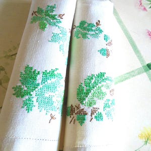 Puede incluir: Dos servilletas de lino blanco enrolladas con dise&ntilde;os de hojas de uva bordadas en verde y marr&oacute;n. Las servilletas est&aacute;n dobladas, mostrando el detallado bordado de punto de cruz. Cada servilleta mide unos 30 cm.