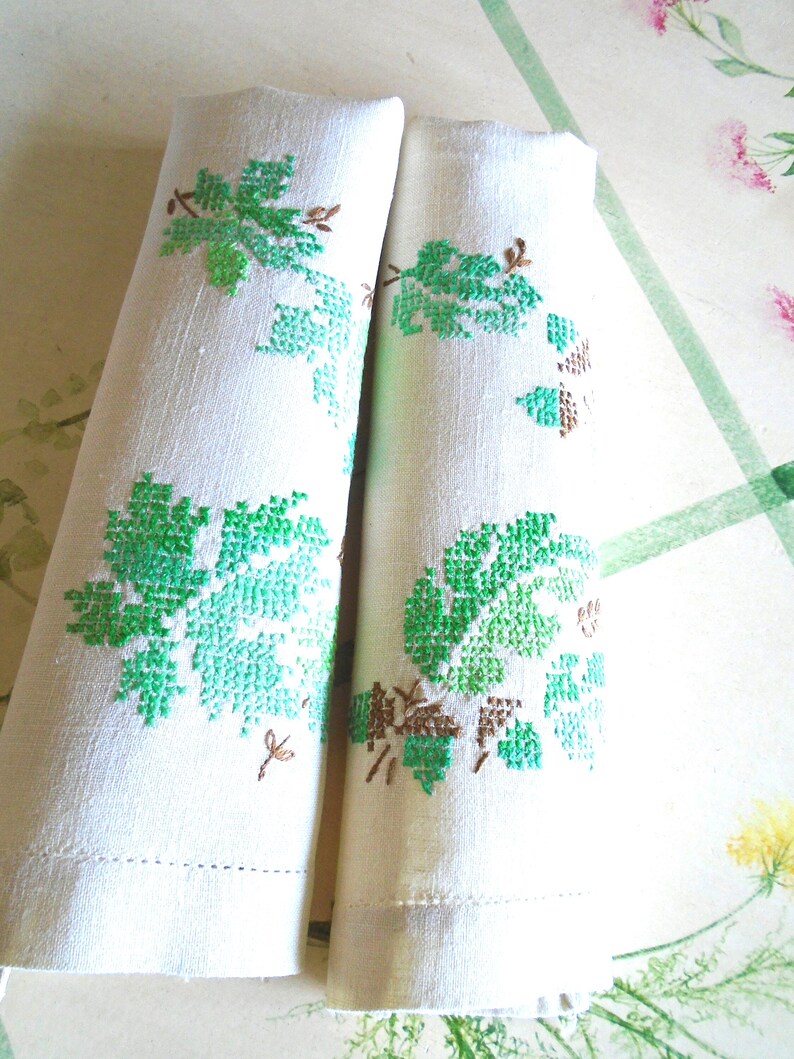 Puede incluir: Dos servilletas de lino blanco enrolladas con dise&ntilde;os de hojas de uva bordadas en verde y marr&oacute;n. Las servilletas est&aacute;n dobladas, mostrando el detallado bordado de punto de cruz. Cada servilleta mide unos 30 cm.