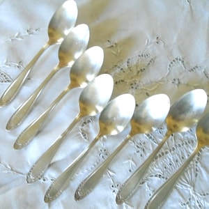 Puede incluir: Una colección de ocho cucharas de plata dispuestas sobre un mantel blanco bordado. Las cucharas tienen un diseño clásico con cuencos alargados y mangos detallados. Las cucharas son reflectantes y el mantel tiene un delicado estampado floral.