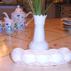 Puede incluir: Un juego de té de porcelana blanca con diseños florales azules, que incluye una tetera, una jarra de crema y un azucarero. Un jarrón blanco con tallos verdes y una fuente en forma de concha también están sobre la mesa. Una pila de platos es visible.