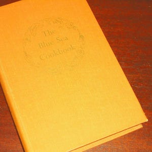 May include: A hardback book with a solid yellow cover featuring the title The Blue Sea Cookbook embossed in the centre. The text is surrounded by a circular decorative border. The book has a clean, uniform appearance with a textured finish.