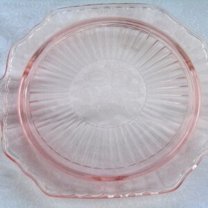 Puede incluir: Un plato de servir de cristal rosa vintage con borde festoneado y dise&ntilde;o radial. El plato tiene un centro circular con un motivo floral y dos asas peque&ntilde;as en lados opuestos. El plato es de cristal transparente.