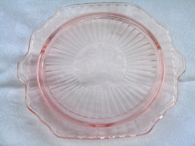 Puede incluir: Un plato de servir de cristal rosa vintage con borde festoneado y dise&ntilde;o radial. El plato tiene un centro circular con un motivo floral y dos asas peque&ntilde;as en lados opuestos. El plato es de cristal transparente.