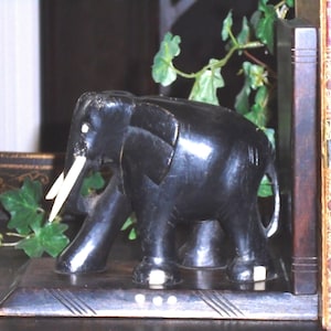 Puede incluir: Un sujetalibros de elefante tallado en negro con colmillos blancos y pequeños detalles blancos en los pies y la base. El elefante se apoya sobre una plataforma rectangular de madera con líneas grabadas, junto a un libro antiguo con letras doradas.