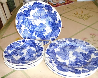 Puede incluir: Un conjunto de platos y un cuenco de cerámica blanca con un diseño de uvas y hojas azules. Los platos están apilados y el cuenco está sobre un soporte dorado. El diseño presenta racimos de uvas y hojas detalladas.