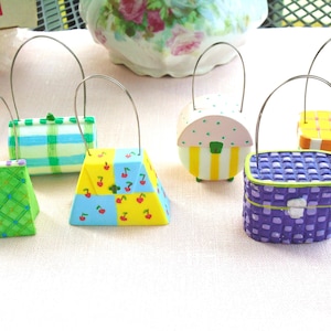 May include: A collection of miniature, decorative purses with wire handles. The purses feature various designs, including plaid, cherries, and woven patterns. Colors include green, blue, yellow, and purple. The purses are displayed on a light pink surface.