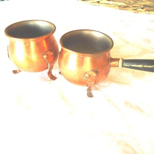 Op de afbeelding: Twee kleine, koperkleurige potten met afgeronde lichamen en drie kleine voetjes. Eén pot heeft een zwarte handgreep. De potten hebben een glanzend, reflecterend oppervlak. De potten zijn waarschijnlijk om te koken of te serveren.