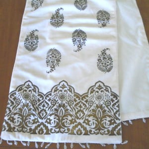 May include: White table runner with a gold and black paisley pattern and a decorative border. The runner has a fringe trim at the bottom. The paisley design is repeated across the fabric.