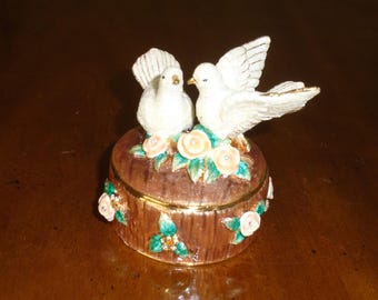 May include: A round brown trinket box with a textured tree bark finish, featuring two white doves perched on top among light pink flowers and green leaves. The sides of the box are decorated with small pink floral accents and green leaves.