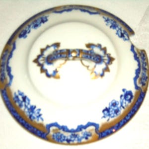 May include: A white porcelain plate with a blue floral design and gold trim. The plate has a central design with a blue and gold accent. The rim features a repeating blue floral pattern. The plate has a small notch on the side.