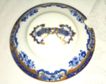 Puede incluir: Un plato de porcelana blanca con un diseño floral azul y ribete dorado. El plato tiene un diseño central con un acento azul y dorado. El borde presenta un patrón floral azul repetido. El plato tiene una pequeña muesca en el lateral.