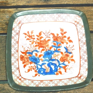 Puede incluir: Un plato cuadrado de cerámica con un borde de metal gris oscuro. El centro presenta un fondo blanco con un diseño floral pintado a mano en naranja y azul vibrantes. Un patrón de cuadrícula rojo bordea el borde interior de la superficie.