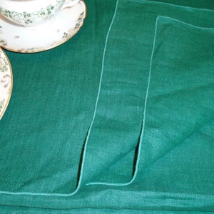 Puede incluir: Una servilleta de lino verde doblada con una textura sutil. La servilleta está doblada en tres y tiene un borde ligeramente deshilachado.