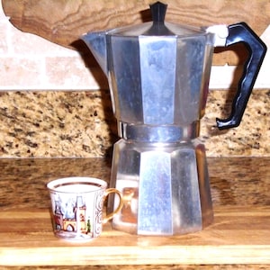 May include: A silver stovetop espresso maker with a black handle and a small decorative cup with a gold handle and a castle design. The coffee maker and cup are on a wooden chopping board.