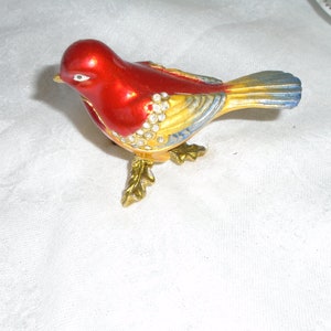 May include: A red, yellow, and blue jeweled bird figurine with a shiny finish. The bird has a long tail and is standing on a gold base.