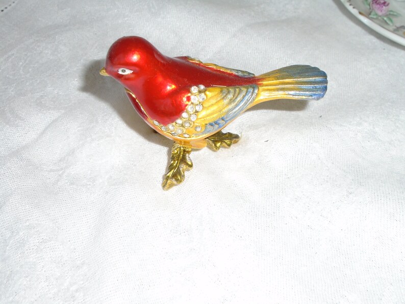 May include: A red, yellow, and blue jeweled bird figurine with a shiny finish. The bird has a long tail and is standing on a gold base.