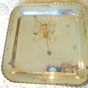 Puede incluir: Una bandeja cuadrada de metal plateado pulido con esquinas redondeadas y un borde festoneado decorativo. El centro de la bandeja presenta un patrón floral grabado y la superficie reflectante muestra el reflejo de una lámpara de techo.