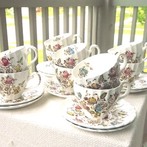 Puede incluir: Una colección de tazas de té de porcelana blanca con platos a juego, que presentan un patrón floral detallado con flores rosas, amarillas y azules y hojas verdes. Las tazas tienen una textura acanalada y asas curvas, dispuestas en pilas.
