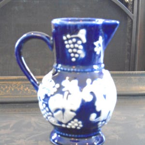 May include: A cobalt blue ceramic pitcher with a handle and spout. The pitcher is decorated with white grape and leaf designs. The handle is curved, and the base is round. The pitcher is likely used for serving beverages.