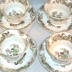 Może przedstawiać: Cztery filiżanki i spodki do herbaty w stylu vintage. Filiżanki i spodki są białe z kwiatowym i przewijanym wzorem w kolorze brązowym, zielonym, żółtym i różowym. Filiżanki do herbaty mają złoty brzeg. Klasyczny zestaw na przyjęcia herbaciane lub do ekspozycji.