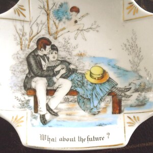 May include: A white porcelain dish with a romantic scene. A couple sits on a bench, with a hat and a cherub in the background. The dish has gold accents and the text "What about the future?"