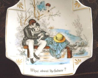 May include: A square white porcelain dish with gold-trimmed corners featuring a colorful illustration of a couple sitting on a bench with a winged figure above them. The text "What about the future?" is printed in black at the bottom of the dish.