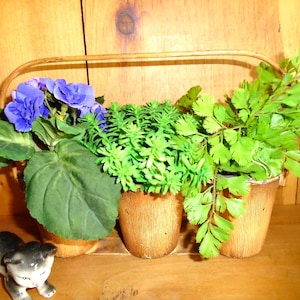 Puede incluir: Una cesta decorativa con tres macetas de madera pequeñas llenas de plantas artificiales. Las plantas incluyen helechos verdes, suculentas y flores moradas. Una pequeña figura de gatito de cerámica en blanco y negro está frente a la cesta.
