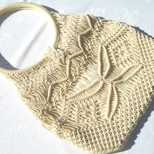 May include: Cream-colored macrame tote bag with a circular handle. The bag features a floral design in the center and a woven pattern throughout. The bag is made of a soft, woven material.