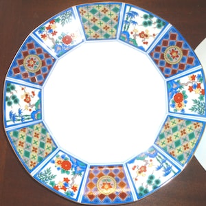 May include: Decorative ceramic plate with a white center and a patterned border. The border features a repeating design of floral and geometric patterns in red, blue, green, and yellow. A second white plate is partially visible.