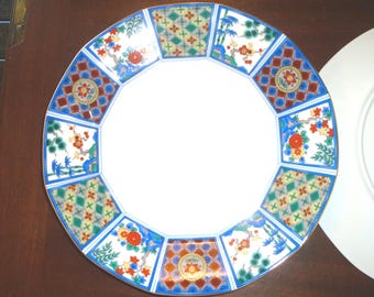 Puede incluir: Un plato redondo de cerámica con un centro blanco y un borde decorativo que presenta paneles alternos de patrones geométricos de diamantes en verde y oro, y motivos florales con acentos rojos, azules y verdes sobre fondo blanco.