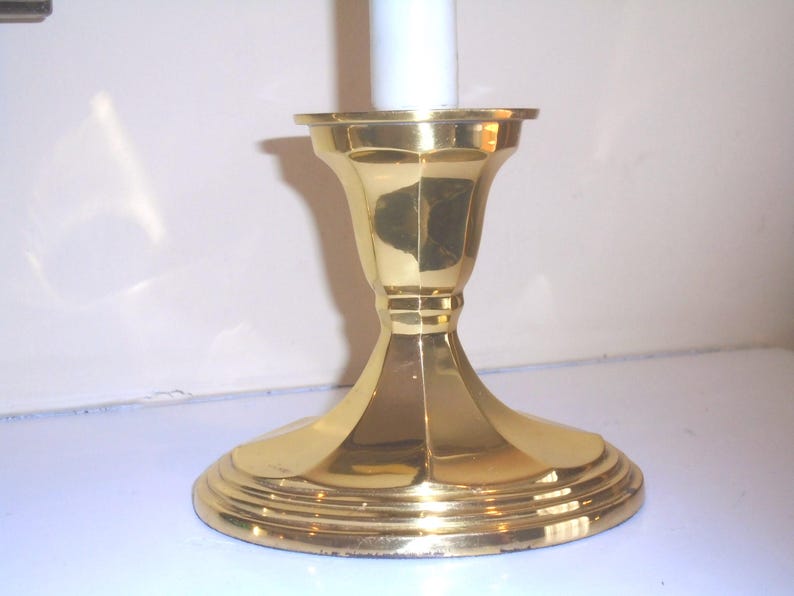 Puede incluir: Un candelero de lat&oacute;n pulido con una base ancha y escalonada y un cuerpo acanalado en forma de campana. El candelero est&aacute; dise&ntilde;ado para sostener una sola vela blanca. El dise&ntilde;o general es cl&aacute;sico y elegante.