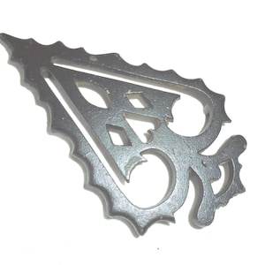 May include: A decorative, gray metal piece with an ornate, openwork design. The piece has a scalloped edge and features a stylized letter "B" with diamond-shaped cutouts. The overall shape is reminiscent of a stylized leaf or emblem.