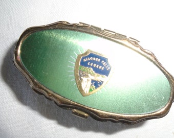 May include: A small, oval-shaped, green metal trinket box with a gold trim. The box has a hinged lid and features a blue and gold enamel emblem with the words "Niagara Falls Canada".