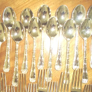 May include: A collection of silver-colored spoons and forks arranged on a wooden surface. The silverware has a decorative floral pattern on the handles. The spoons have a rounded bowl shape, and the forks have four tines.