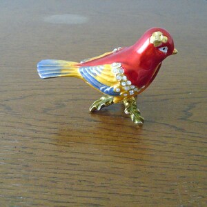 May include: A small, red, yellow, and blue bird figurine with rhinestones. The bird is standing on a gold-colored base with leaves.