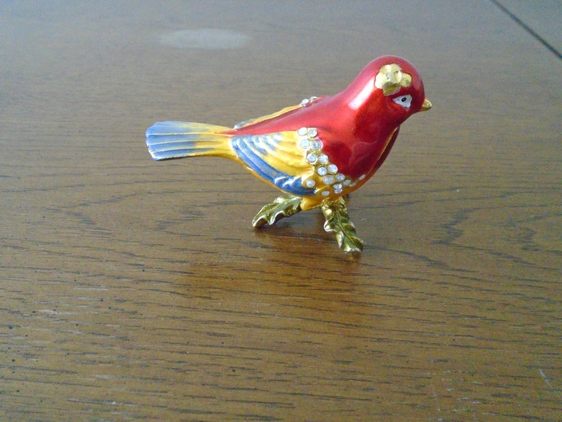 May include: A small, red, yellow, and blue bird figurine with rhinestones. The bird is standing on a gold-colored base with leaves.