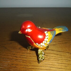 May include: A small, red, yellow, and blue bird figurine with gold legs and rhinestones. The bird is perched on a wooden surface.