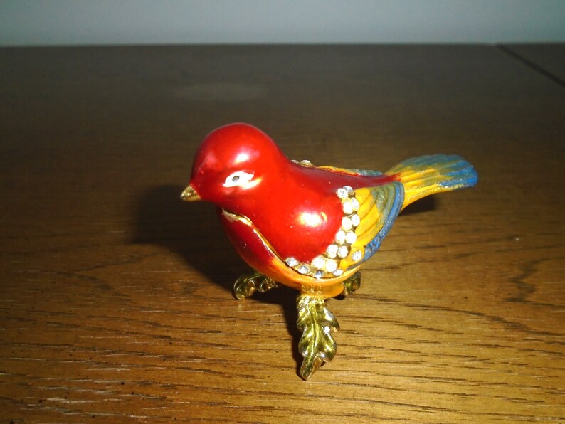 May include: A small, red, yellow, and blue bird figurine with gold legs and rhinestones. The bird is perched on a wooden surface.