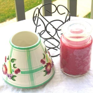 May include: A candle in a clear glass jar with red wax and a glass lid. A white and green lampshade with fruit and green stripe designs sits next to a black metal wine rack.