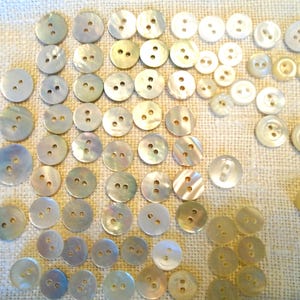 May include: A collection of various sizes of iridescent, pearlescent buttons with two or four holes. The buttons are arranged on a light beige fabric, showcasing their subtle color variations and textures. Ideal for sewing and craft projects.