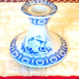Puede incluir: Un candelabro de cerámica blanca con un patrón azul de estilo transferware. El diseño incluye una escena paisajística arquitectónica central rodeada por un borde decorativo de motivos repetitivos en forma de concha en la base y el borde.