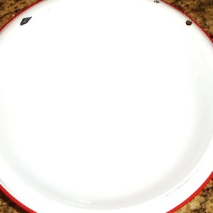 Puede incluir: Un plato de esmalte blanco con borde rojo. El plato tiene una forma ligeramente cóncava y muestra algunos signos de desgaste. Este plato de estilo vintage es adecuado para servir alimentos.