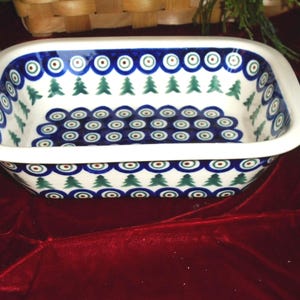 May include: Rectangular ceramic baking dish with a white base, blue rim, and a repeating pattern of green fir trees and blue circles. The dish has a white rim and is likely used for baking or serving.