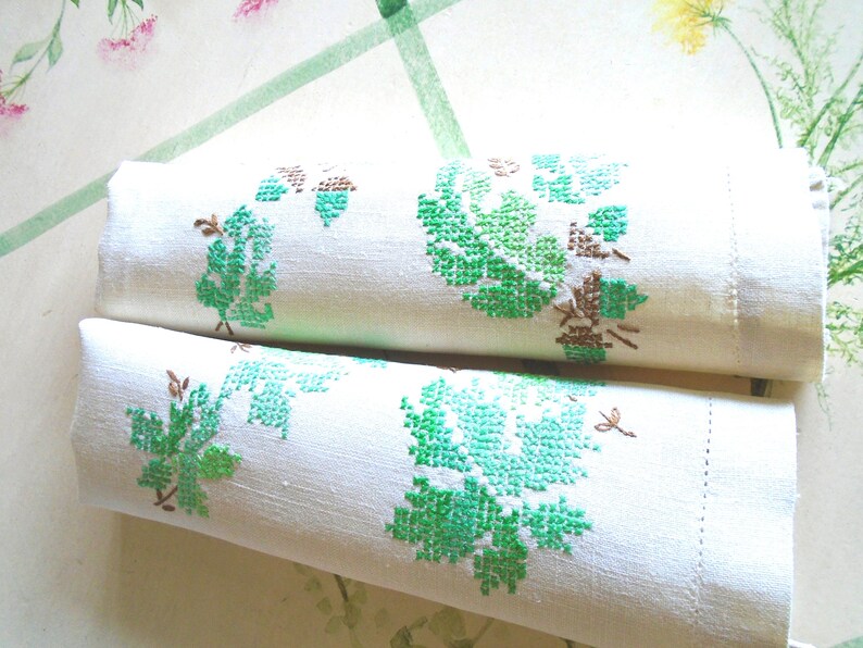 Puede incluir: Dos toallas de lino blanco enrolladas con bordado floral verde y marr&oacute;n. El bordado presenta un dise&ntilde;o detallado de hojas y vides. Las toallas est&aacute;n cuidadosamente enrolladas, mostrando la intrincada labor.