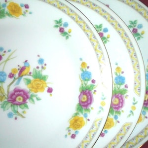 May include: A set of three white porcelain plates with a floral and bird design. The plates feature a colorful pattern of yellow, purple, and blue flowers, with a small bird perched on a branch. A gold trim and a decorative border complete the design.