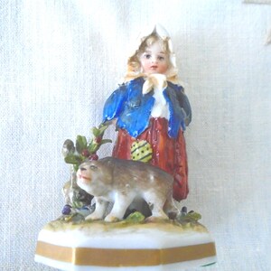 Puede incluir: Una figura de porcelana que representa a un niño con un gorro blanco, una chaqueta azul y una falda roja, de pie junto a un perro marrón. La figura se asienta sobre una base hexagonal blanca con una franja dorada y detalles de follaje.