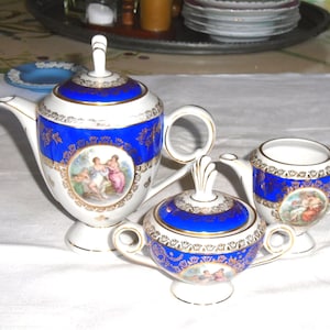 May include: A white and blue porcelain tea set with gold trim. The set includes a teapot, sugar bowl, and creamer. Each piece features a blue band with gold detailing and a painted scene of figures.