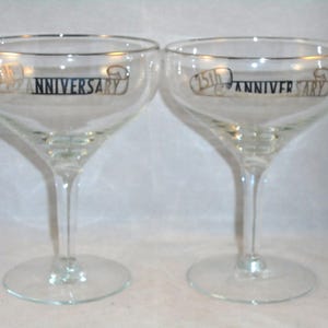 Puede incluir: Dos copas de champán transparentes con un cuenco ancho y un tallo delgado. Las copas tienen un diseño dorado y negro que dice "75th ANNIVERSARY". Las copas están sobre un fondo blanco.
