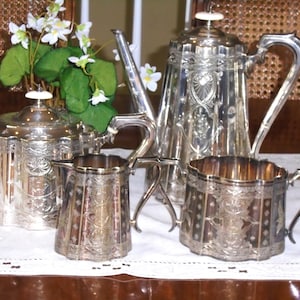 May include: A silver tea service set consisting of a tall coffee pot, a shorter teapot, a creamer, and a sugar bowl. Each piece features intricate embossed floral and geometric patterns, scalloped edges, and polished silver surfaces with handles.