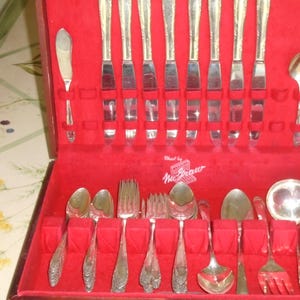 May include: A vintage silverware set in a red velvet-lined case. The set includes knives, forks, spoons, and serving utensils. The silver-toned flatware has decorative handles. The case is marked "West by McGraw".